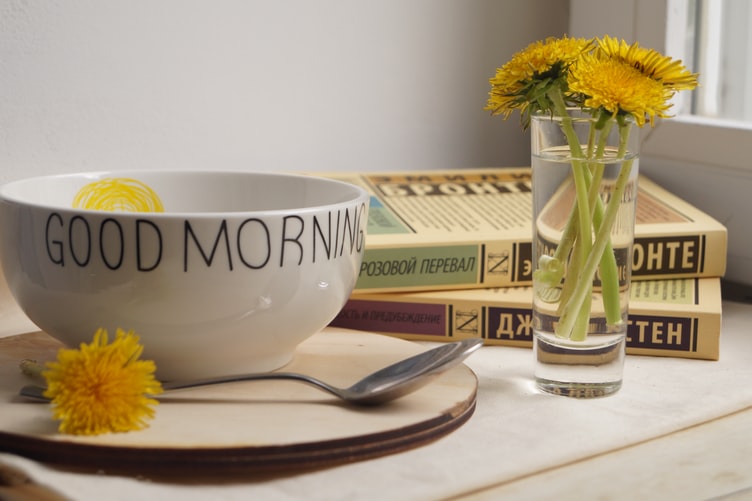A bowl, plate, spoon, a few books and a small glass of water with yellow flowers on a table