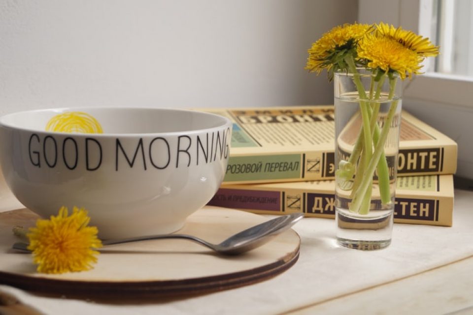 A bowl, plate, spoon, a few books and a small glass of water with yellow flowers on a table