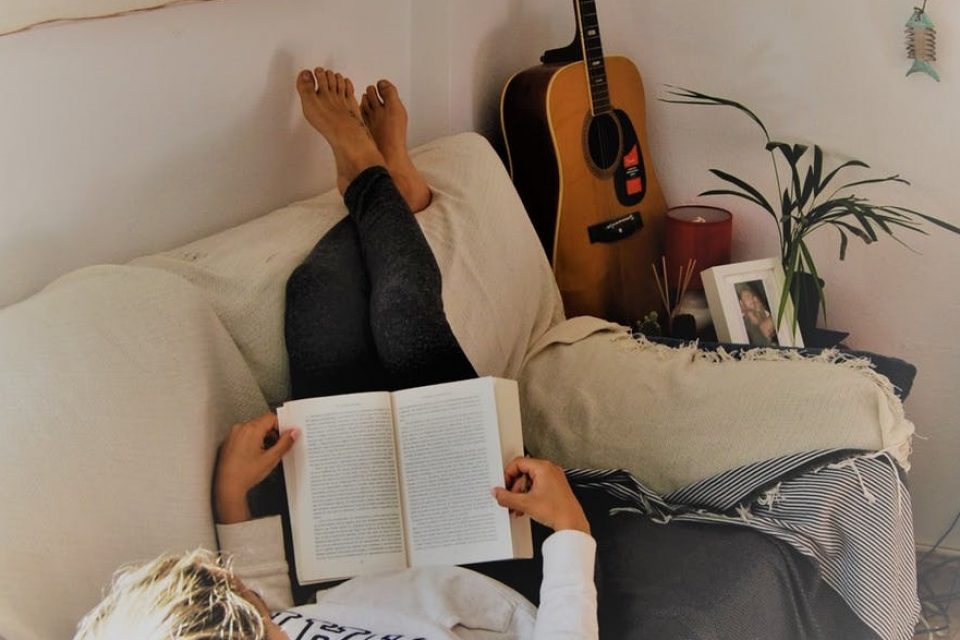 A woman laying on her back on the sofa with her legs up resting on the top of the sofa, reading a book